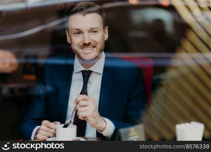 Horizontal shot of happy businessman dressed in formal clothing, drinks aromatic hot beverage, looks at window while sits in cafeteria, has pleased expression. People, rest and lifestyle concept
