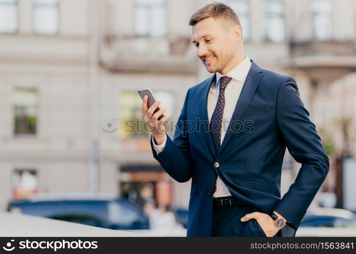 Horizontal shot of happy businessman keeps hand in pocket, wears formal suit and wristwatch, recieves notification on smart phone, finds out about high financial balance. Cheerful trader outdoor