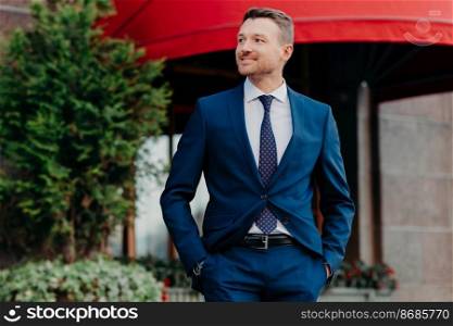 Horizontal shot of happy businessman looks cheerfully and confidently aside, keeps hands in pockets, poses outdoor, waits for business partner. Male banker has dinner break, rests outside alone
