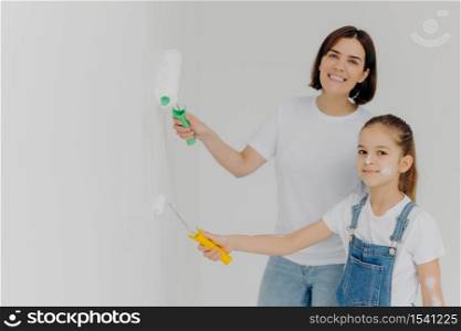 Horizontal shot of happy girl and her mother paint walls in white color, use paint rollers, improve house, busy with domestic work, move in new apartment. Home repair and improvement concept.
