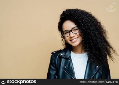 Horizontal shot of happy healthy woman with natural curly hair, pleasant smile, enjoys spare time and doing shopping at weekend, wears transparent glasses, leather jacket, isolated on beige background