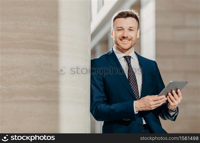 Horizontal shot of happy male manager dressed in black formal suit, uses modern tablet computer for making financial report, focused into distance with happy expression. People and business concept