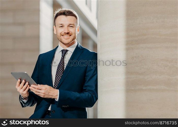 Horizontal shot of happy male manager dressed in black formal suit, uses modern tablet computer for making financial report, focused into distance with happy expression. People and business concept