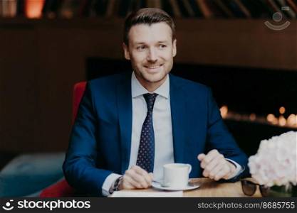 Horizontal shot of happy male office worker has coffee break at cafeteria, drinks hot beverage, dressed in formal clothing, sits in restaurant, looks joyfully into distance, focused somewhere