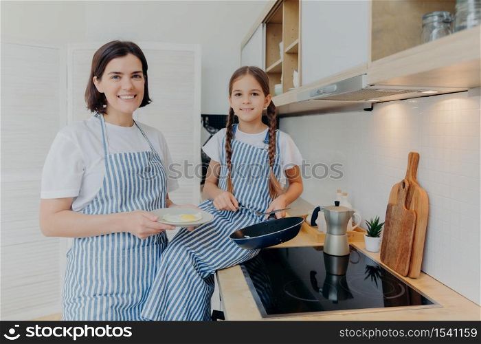 Horizontal shot of happy mother and small female child look gladfully at camera, busy preparing fried eggs for breakfast or dinner, pose together near cooker, being at kitchen. Family concept