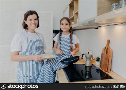 Horizontal shot of happy mother and small female child look gladfully at camera, busy preparing fried eggs for breakfast or dinner, pose together near cooker, being at kitchen. Family concept