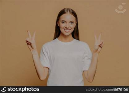 Horizontal shot of happy optimistic young Caucasian woman raises arms shows peace gesture with fingers does victory sign says number two wears casual white t shirt isolated over brown background