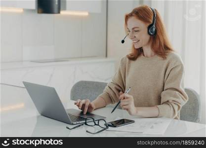 Horizontal shot of happy redhead woman watches online webinar participates in online course uses headset focused at laptop screen holds pen makes notes surrounded by documents communicates online. Happy redhead woman watches online webinar uses headset focused at laptop screen
