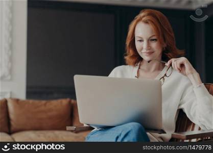 Horizontal shot of happy skilled ginger woman teacher works distantly, checks students works via laptop, holds spectacles, poses in modern cozy apartment. Student checks database for university