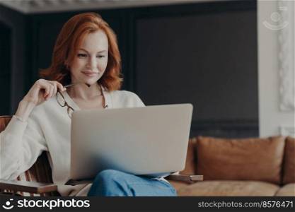 Horizontal shot of happy skilled ginger woman teacher works distantly, checks students works via laptop, holds spectacles, poses in modern cozy apartment. Student checks database for university