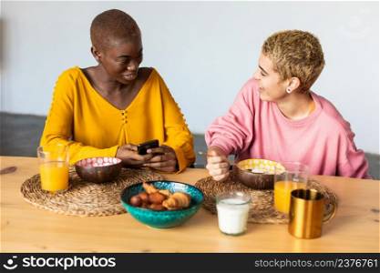 Horizontal shot of happy women discuss funny blog in internet, use mobile phone, eating healthy dish at home