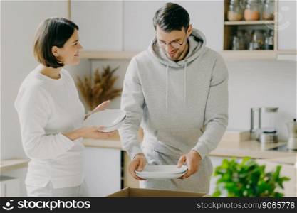 Horizontal shot of happy young woman and man open boxes with dishes, placing plates in modern kitchen, relocate in≠w housing, start≠w life in modern apartment, rent house for living to≥ther