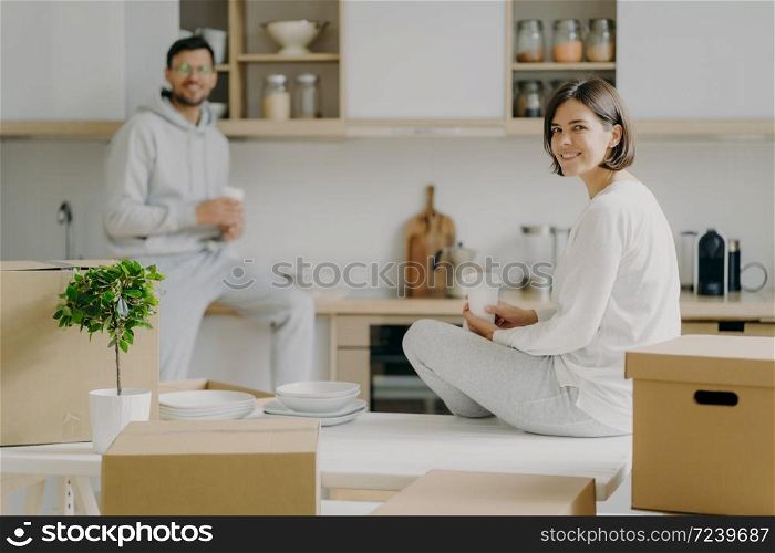 Horizontal shot of happy young woman and man sit on kitchen desk, drink coffee, have break after unpacking boxes, have pleasant talk, surrounded with plates, indoor plant in pot. Moving concept