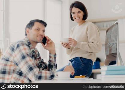 Horizontal shot of husband and wife analyze utilities bills, study bank loan documents, manage finances and plan expenses, make telephone call, woman drinks coffee and ready to give useful advice