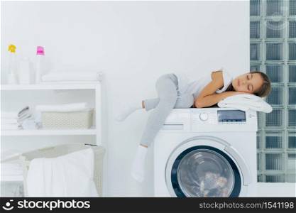 Horizontal shot of little girl has rest on washing machine, feels tired after helping mum about house, sleeps on washer poses in laundry room with console, detergents, linen around. Household routine