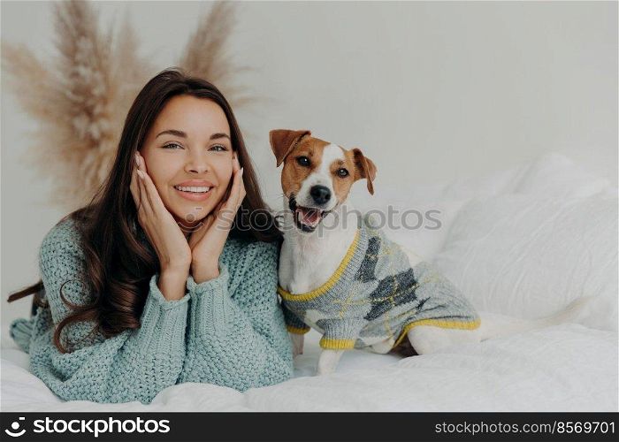 Horizontal shot of lovely brunette female in casual wear, lies on white bedclothes with pedigree dog, expresses happiness, enjoys company of pet, rest together at home, feel relaxed and satisfied