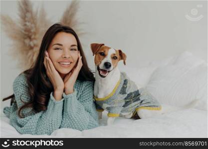 Horizontal shot of lovely brunette female in casual wear, lies on white bedclothes with pedigree dog, expresses happiness, enjoys company of pet, rest together at home, feel relaxed and satisfied