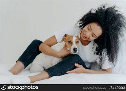 Horizontal shot of lovely curly woman embraces favourite dog, wears whihte t shirt, jeans and socks, enjoy time together, sit on bed. Good beginning of day. Cheerfull lady plays with pet in bedroom