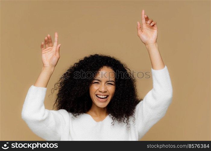Horizontal shot of lovely dark skinned female model has toothy smile, raises hands, points up, feels overjoyed, has curly hair, wears white sweater, isolated over brown background. Woman has fun