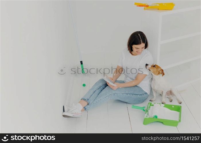 Horizontal shot of lovely woman with short hair, looks attentively at color palette, chooses colour for painting room, plays with pedigree dog, sit in white spacious room, sits on white floor