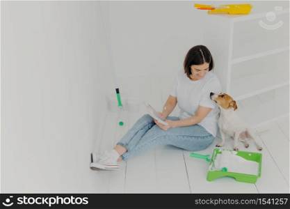 Horizontal shot of lovely woman with short hair, looks attentively at color palette, chooses colour for painting room, plays with pedigree dog, sit in white spacious room, sits on white floor