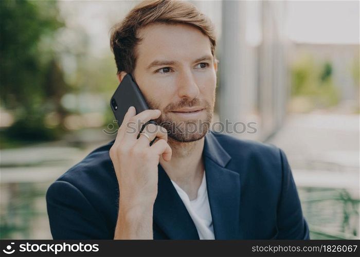 Horizontal shot of male entrepreneur or manager consults client by telephone call discusses future project has self confident expression poses outdoor wears formal clothes has busy working day. Horizontal shot of male entrepreneur or manager consults client by telephone call