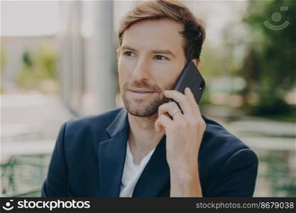 Horizontal shot of male entrepreneur or manager consults client by telephone call discusses future project has self confident expression poses outdoor wears formal clothes has busy working day. Horizontal shot of male entrepreneur or manager consults client by telephone call