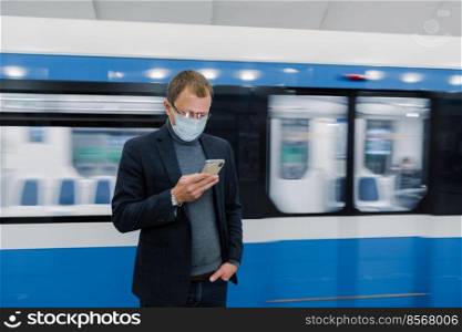 Horizontal shot of male worker poses at subway platform, commutes by public transport, uses modern mobile phone to check route, wears medical protective mask against coronavirus or flu. Health hazard