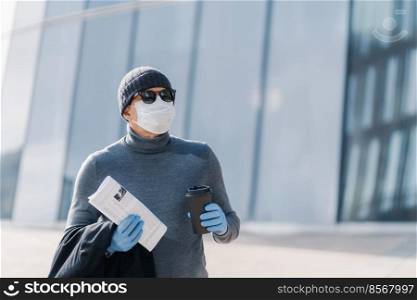 Horizontal shot of man being in danger to catch coronavirus in public place, protects himself by wearing medical mask and rubber gloves, drinks takeaway coffee, looks somewhere through sunglasses