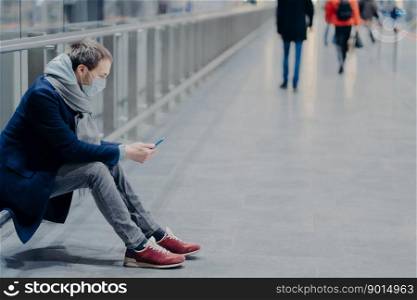 Horizontal shot of man holds modern mobile phone, being in mall during pandemic and coronavirus, wears protective medical mask, dressed casually, checks mobile application. Health care concept