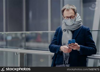 Horizontal shot of man in spectacles uses modern mobile phone, wears medical mask during coronavirus outbreak, has influenza protection in big shopping mall. Flu and corona season. Health care concept