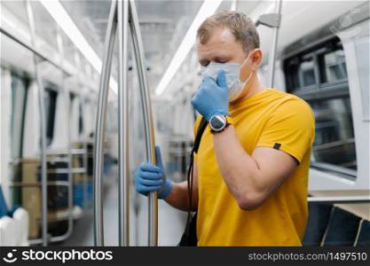 Horizontal shot of man passenger coughs and has problems with breathing, wears disposable mask and gloves, stands in public transport, metro, prevents from coronavirus. Public health solution