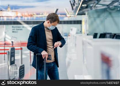 Horizontal shot of man passenger wears disposable medical mask to prevent virus, checks time on his watch, has hour for departure, poses in internation airport with suitcase and passport. Coronavirus