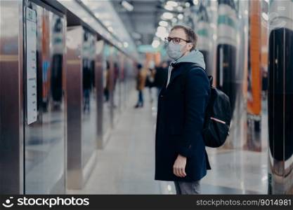 Horizontal shot of man traveler poses on metro station, wears medical mask during coronavirus time, waits for train, commutes to work by public transport. Virus, disease and influenza concept
