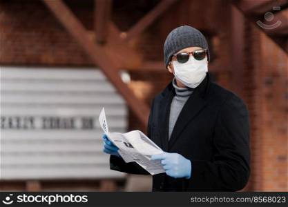 Horizontal shot of man uses virus preventive measures during coronavirus pandemic, wears face medical mask and rubber gloves, reads newspaper. Epidemic disease, Covid-19, health care concept