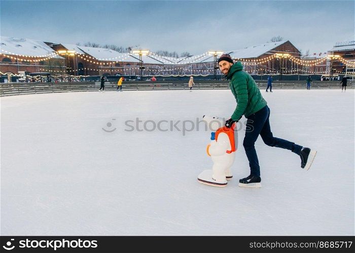 Horizontal shot of man wears gree anorak, warm hat and skates,≤arns to go skating on ice, uses skate aid, enjoys free time and w∫er holidays, smi≤s hapπly at camera. Hobby and peop≤concept