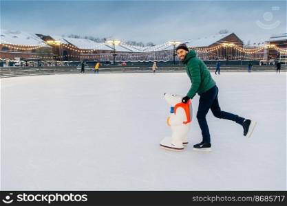 Horizontal shot of man wears gree anorak, warm hat and skates,≤arns to go skating on ice, uses skate aid, enjoys free time and w∫er holidays, smi≤s hapπly at camera. Hobby and peop≤concept