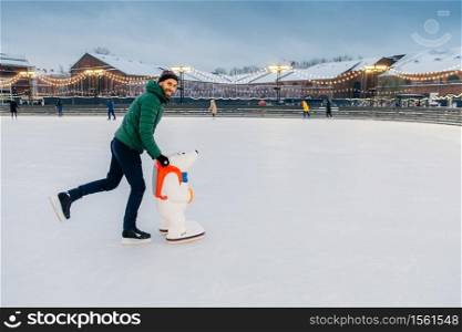 Horizontal shot of man wears gree anorak, warm hat and skates, learns to go skating on ice, uses skate aid, enjoys free time and winter holidays, smiles happily at camera. Hobby and people concept