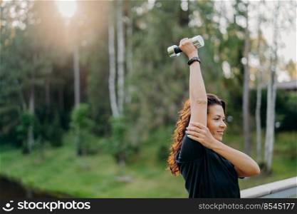 Horizontal shot of motivated brunette woman raises arm and holds dumbbell, trains muscles, has regular morning workout, wears black t shirt, poses outdoor against trees background. Bodybuilding