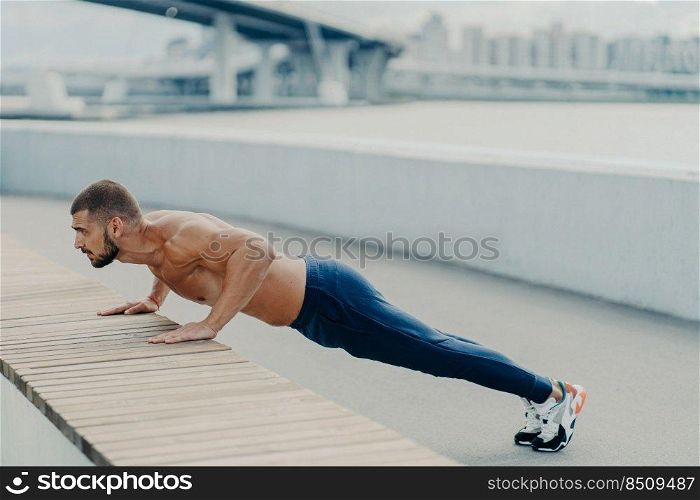 Horizontal shot of muscular athlete man does push up exercise has strong body stands in plank pose outdoor wears sport trousers and sneakers concentrated into distance. Strength and motivation concept