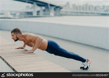 Horizontal shot of muscular athlete man does push up exercise has strong body stands in plank pose outdoor wears sport trousers and sneakers concentrated into distance. Strength and motivation concept