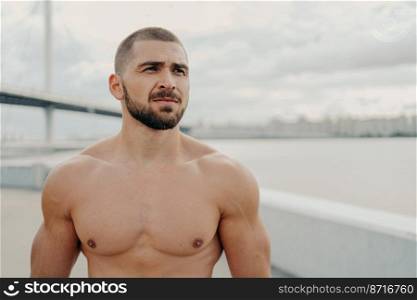 Horizontal shot of muscular man with naked torso thick beard looks somewhere into distance, relaxes outdoors, stands against river background, takes break after jogging, has athletic body shape