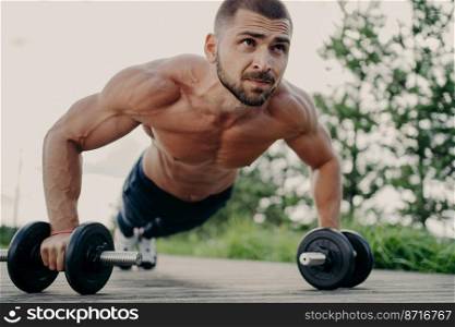 Horizontal shot of muscular unshaven man does push up exercises, leans at barbells, focused into distance, puts all efforts while trains abdominal muscles poses outdoor. Sporty lifestyle concept