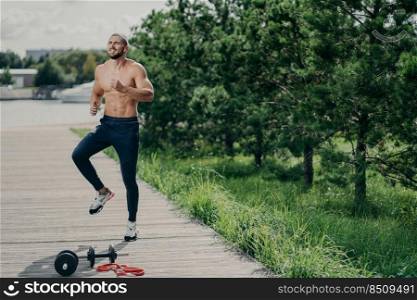 Horizontal shot of naked active man with naked torso jumps on one place, does sport exercises, poses outdoor, wears sport trousers and sneakers, warms up before jogging. Active lifestyle concept
