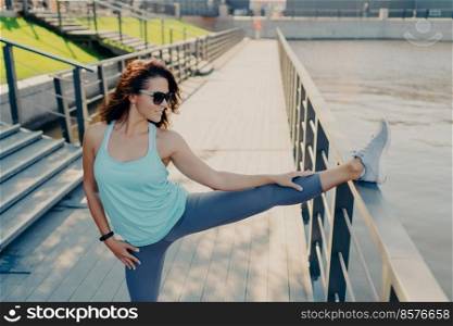 Horizontal shot of of brunette woman wears casual sport clothes stretches legs on fence has athletic body warms up before cardio training has flexible fit arms and legs poses outside. Active lifestyle