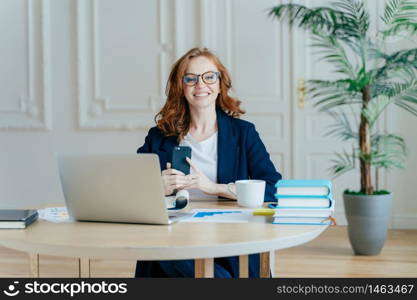 Horizontal shot of optimistic woman installs application on modern cell phone, checks notification, surrounded with paper documents, works on laptop computer, has red hair, dressed formally.