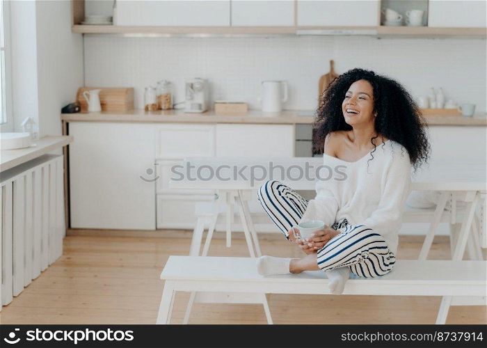 Horizontal shot of overjoyed dark skinned woman laughs pleasantly, drinks coffee, looks out of window in kitchen, dressed in fashionable clothes. Smiling lady with hot tasty beverage, relaxes at home