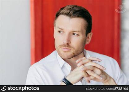Horizontal shot of pensive handsome young male corporate worker holds hands together, looks aside, being deep in thoughts after meeting, poses over red background. Man leader owns big company