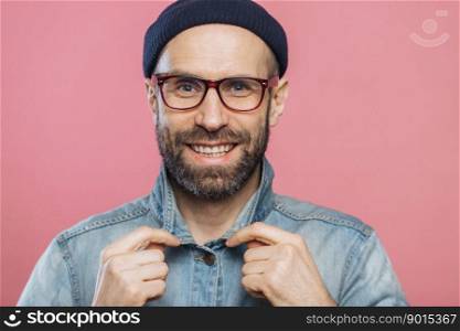 Horizontal shot of pleasant looking smiling bearded male wears glasses and denim jacket, advertizes new fashionable clothes, has happy expression, isolated over pink background. Positive emotions
