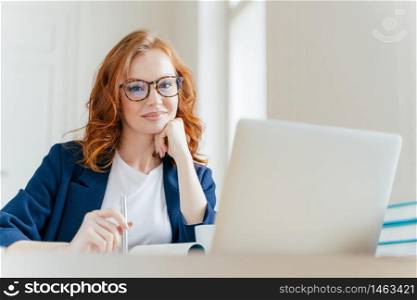 Horizontal shot of pleasant looking successful professional female lawyer learns clients case, works on modern laptop computer, dressed in formal apparel and transparent glasses, poses in office
